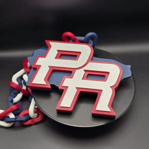 Puede incluir: Un emblema 3D de Puerto Rico con las letras "PR" en blanco con borde rojo, sobre un fondo azul con forma de isla. Una cadena en rojo, blanco y azul está adjunta.