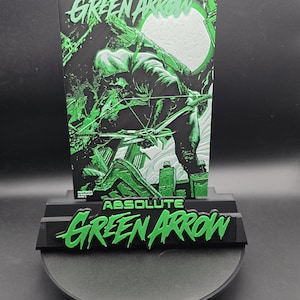 May include: A black and green display stand featuring a vertical plaque with the text ABSOLUTE GREEN ARROW and a graphic of a hooded archer aiming a bow. The base of the stand also displays the text ABSOLUTE GREEN ARROW in bold green lettering.