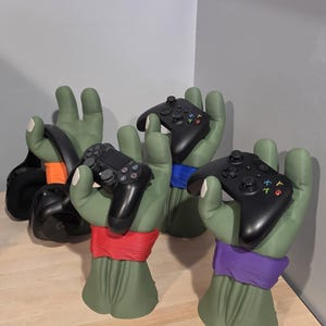 May include: Three green hand-shaped stands with wristbands in red, blue, and purple, each holding a black gaming controller or headset. The stands feature white fingernails and are designed to display gaming accessories on a flat surface.