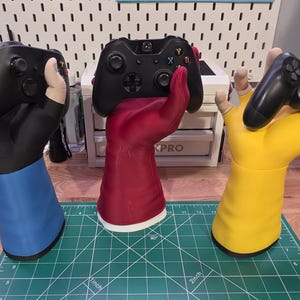 May include: Three upright hand-shaped controller stands in blue, red, and yellow. Each stand features a fingerless glove design and holds a black gaming controller. The stands are positioned on a green cutting mat with visible inch markings.