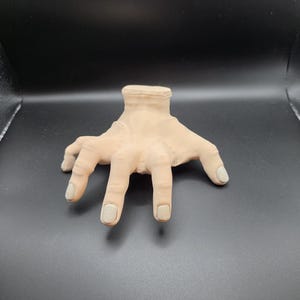 May include: A beige-colored, disembodied hand with gray painted fingernails. The hand is posed with fingers spread and the wrist area is visible. The hand is made of a soft material and is likely a prop or decorative item.