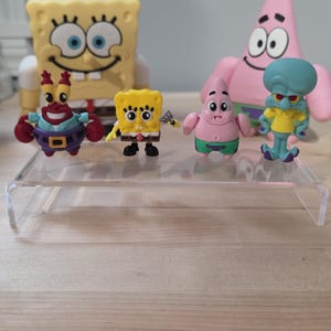 Puede incluir: Una colección de seis figuras de personajes de Bob Esponja. El conjunto incluye dos figuras grandes de Bob y Patricio, y cuatro figuras más pequeñas de Don Cangrejo, Bob con una espátula, Patricio y Calamardo, dispuestas sobre un soporte transparente.
