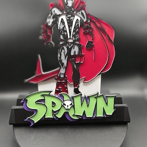 May include: A Spawn figurine, featuring a detailed character design with a red cape and a black and white suit. The base displays the word "SPAWN" in green and purple, with a skull graphic. The figurine is set against a dark background.