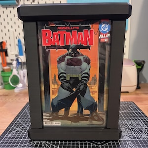 Peut inclure: Un cadre de présentation rectangulaire noir contenant une bande dessinée intitulée Absolute Batman All In 1. La couverture présente une illustration centrale de Batman en costume gris et noir sur fond orange, avec le logo DC en haut à droite.