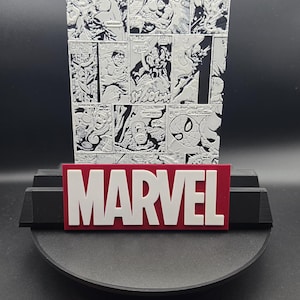 Peut inclure: Un présentoir Marvel avec un arrière-plan de bande dessinée et le logo Marvel rouge et blanc. Le présentoir noir est circulaire, conçu pour exposer des objets de collection ou des souvenirs.