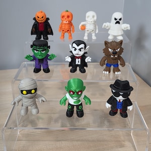 May include: A collection of colorful Halloween-themed figurines, including a pumpkin head, Frankenstein's monster, a vampire, a werewolf, a mummy, and a skeleton. The figures are displayed on a clear, tiered shelf, showcasing their unique designs and vibrant colors.