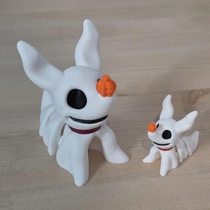 May include: Two white Zero figurines from The Nightmare Before Christmas. The larger figurine has a red collar and an orange pumpkin nose. The smaller figurine has the same features. Both have black eyes and are standing on a light wood surface.