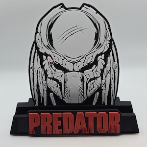 Peut inclure: Un design de masque Predator en noir et blanc avec des accents rouges. Le mot "PREDATOR" est affiché en lettres rouges sur une base noire. Le masque présente des détails et une expression menaçante.