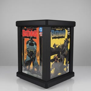Puede incluir: Una vitrina cuadrada negra que contiene dos cómics de Absolute Batman. El lado izquierdo muestra una figura de Batman con traje oscuro sobre un fondo naranja, mientras que el lado derecho presenta un fondo amarillo con una ilustración de acción dinámica.