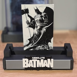 May include: A black and white Batman comic book illustration displayed in a black and gray stand. The stand has the text "ABSOLUTE BATMAN" in white. The comic features Batman holding a weapon.