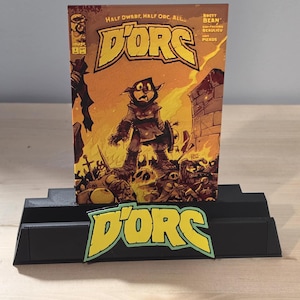 Peut inclure: Une bande dessinée intitulée "D'ORC" avec une illustration fantastique d'un personnage mi-nain, mi-orc. La bande dessinée est présentée sur un présentoir noir à plusieurs niveaux avec le logo "D'ORC" assorti.