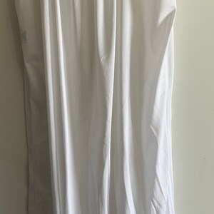 Puede incluir: Un vestido blanco de slip con ribete de encaje alrededor del escote y las sisas. El vestido est&aacute; colgado en una percha de madera.