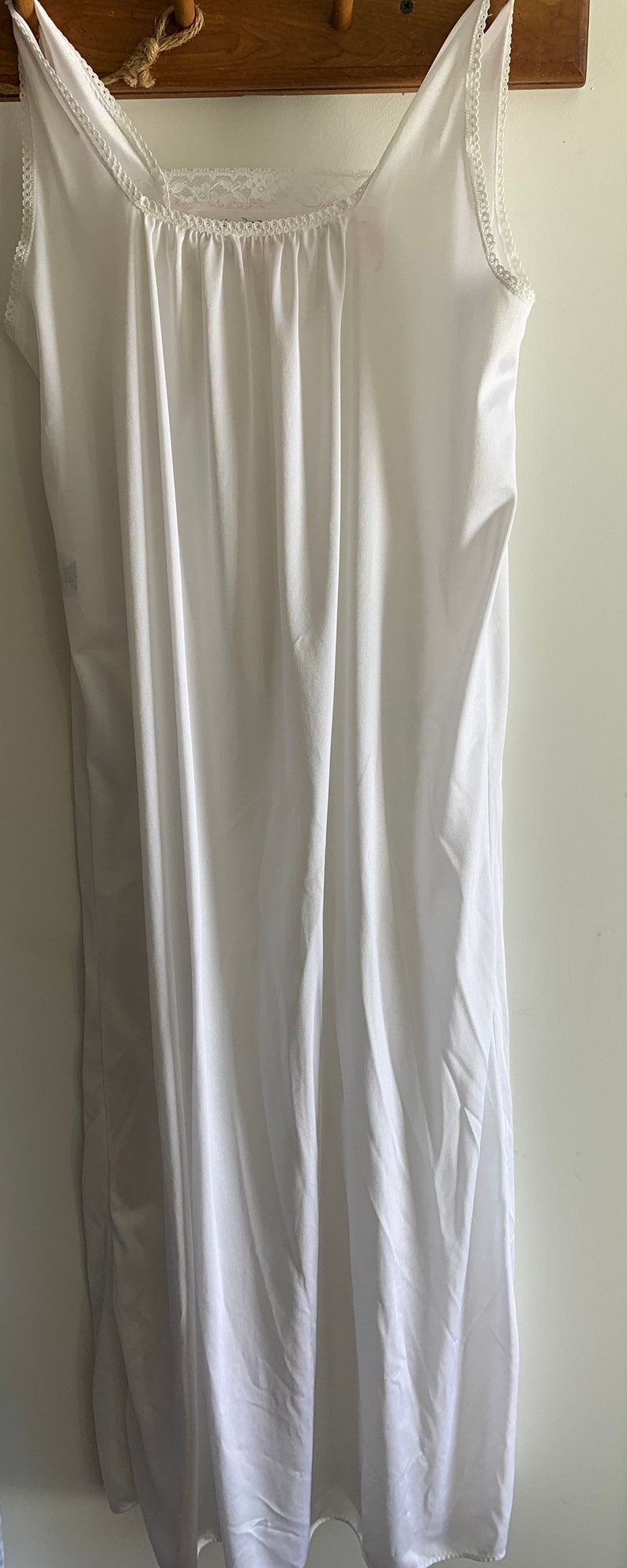 Puede incluir: Un vestido blanco de slip con ribete de encaje alrededor del escote y las sisas. El vestido est&aacute; colgado en una percha de madera.