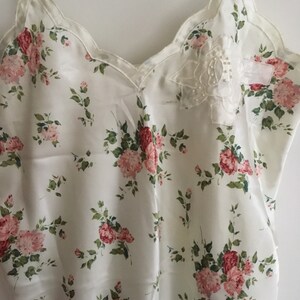 May include: A white satin slip with a floral print of pink roses and green leaves. The neckline is scalloped and features a small white lace applique with pearl-like embellishments.