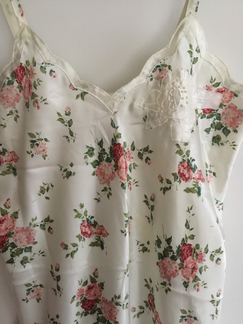 May include: A white satin slip with a floral print of pink roses and green leaves. The neckline is scalloped and features a small white lace applique with pearl-like embellishments.