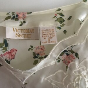 May include: White satin sleep shirt with a floral print and a delicate lace trim. The shirt has a V-neckline and adjustable straps. The tag reads "Victoria's Secret" and includes care instructions.