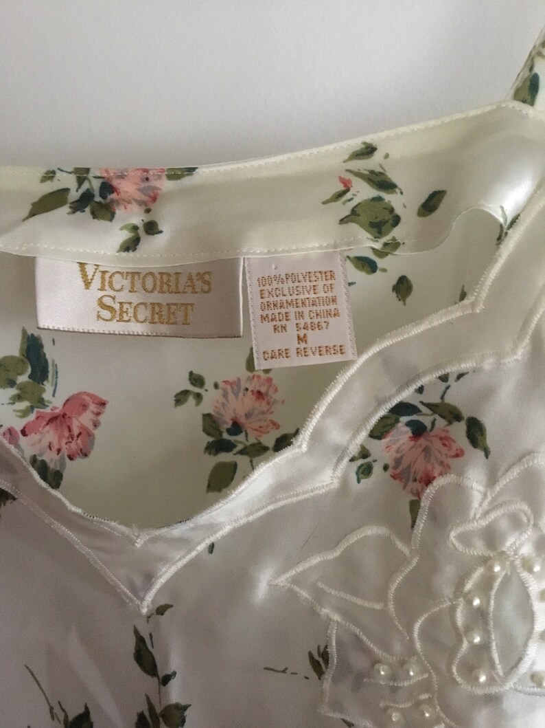 May include: White satin sleep shirt with a floral print and a delicate lace trim. The shirt has a V-neckline and adjustable straps. The tag reads "Victoria's Secret" and includes care instructions.