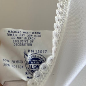 Puede incluir: Una etiqueta de cuidado blanca con texto azul y un logotipo azul y blanco. El texto dice "Machine wash warm, tumble dry low heat, do not bleach, exclusive of decoration, RN 15017, 65% polyester, 35% cotton, ILGWU".
