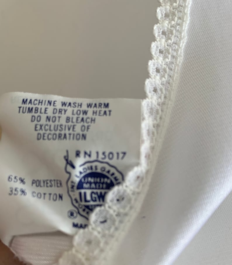 Puede incluir: Una etiqueta de cuidado blanca con texto azul y un logotipo azul y blanco. El texto dice "Machine wash warm, tumble dry low heat, do not bleach, exclusive of decoration, RN 15017, 65% polyester, 35% cotton, ILGWU".