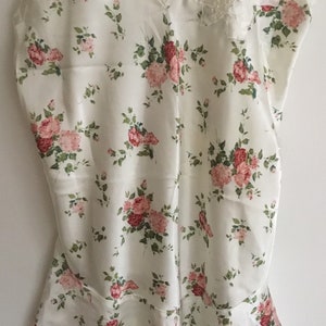 May include: A white satin slip with a floral print featuring pink and red roses. The slip has thin straps and a scalloped neckline. There is a small lace detail on the front of the slip.