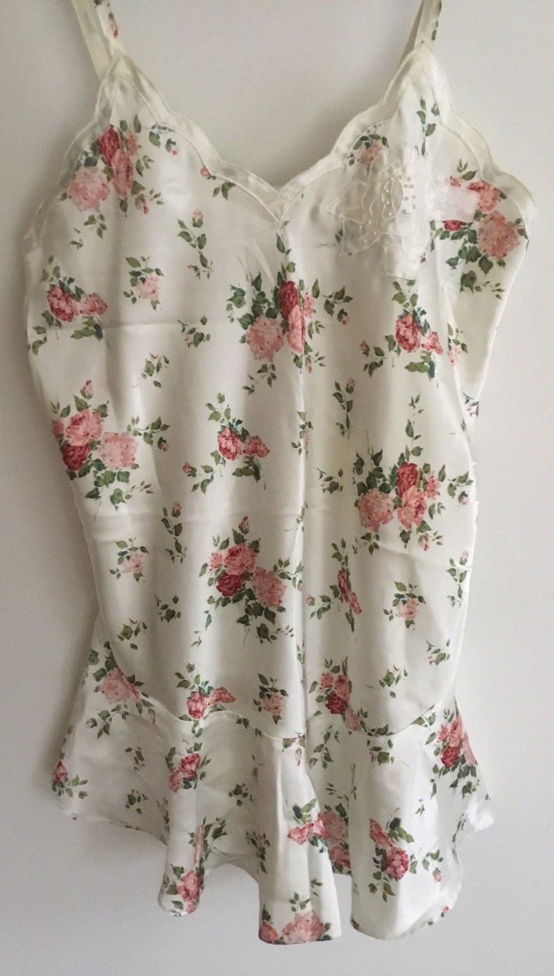 May include: A white satin slip with a floral print featuring pink and red roses. The slip has thin straps and a scalloped neckline. There is a small lace detail on the front of the slip.