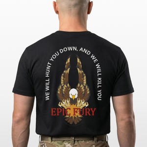 May include: Black t-shirt featuring a detailed eagle graphic with outstretched wings. The design includes the text "WE WILL HUNT YOU DOWN, AND WE WILL KILL YOU" and "EPIC FURY" in red. The shirt is made of a dark fabric.