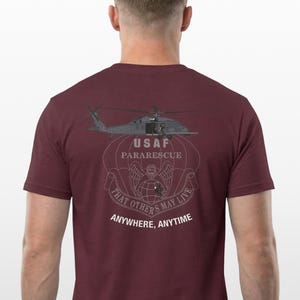 Könnte beinhalten: Burgunderfarbenes T-Shirt mit einem Hubschrauber-Motiv und dem Text "USAF Pararescue". Das Design beinhaltet ein Banner mit den Worten "THAT OTHERS MAY LIVE" und "ANYWHERE, ANYTIME".