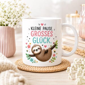 Könnte beinhalten: Eine weiße Keramiktasse mit einem braunen Faultier, das an einem Ast hängt, umgeben von bunten Blumen und Blättern. Der Text "KLEINE PAUSE. GROSSES GLÜCK." ist in Schwarz, Pink und Türkis gedruckt, verziert mit kleinen Herzen und Punkten.