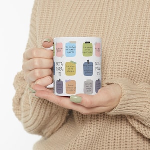 Peut inclure: Mug en céramique blanche avec des boîtes de texte colorées contenant des phrases en espagnol. Le mug est tenu par une personne portant un pull beige. Le texte comprend des phrases comme "Yo activo mi buena suerte".