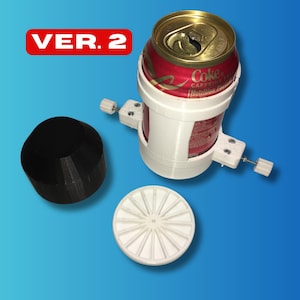 May include: A white 3D-printed can holder with adjustable arms, holding a red and white Coke can. A black, geometric object and a white, circular object with radial lines are also present. The text "VER. 2" is in a red rectangle.