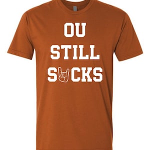 May include: A burnt orange t-shirt with the white text "OU STILL SUCKS" and a hand gesture graphic. The short-sleeved shirt is made of a soft material.