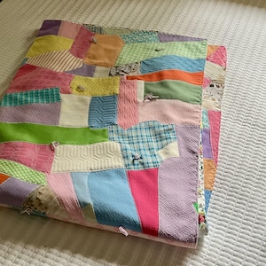 May include: A patchwork quilt constructed from various fabric scraps in a multitude of colors including yellow, pink, blue, green, and orange. The quilt features diverse textures and patterns, such as plaid, floral, and solid weaves, joined together with visible stitching and small tied knots throughout the surface.