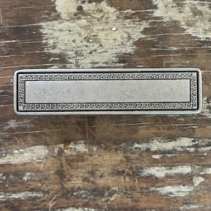 May include: A rectangular silver-toned metal bar with a smooth, flat center panel and a decorative border featuring a continuous black Greek key pattern. The object has a metallic finish with subtle signs of wear and a uniform, elongated shape.