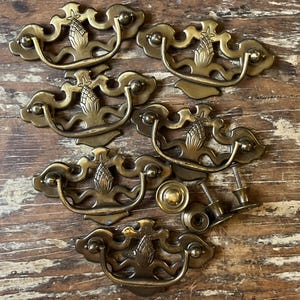 May include: A collection of six brass drawer pulls featuring a central pineapple motif on the backplate and a curved swing handle. The set includes several small metal mounting screws and circular hardware components for installation.