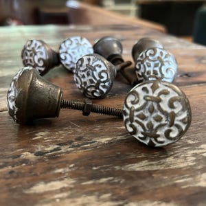 May include: A collection of six cabinet knobs featuring a round, white ceramic face with a raised, dark bronze-colored metal filigree pattern. Each knob has a dark bronze metal base and a threaded metal screw with a small nut attached for installation.