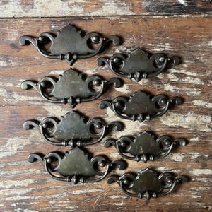 May include: A collection of eight dark bronze-toned metal drawer pulls featuring a decorative backplate with a central crest and curved arms. Each pull includes a swinging bail handle attached to the backplate, showcasing a weathered finish.