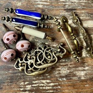 May include: A collection of cabinet hardware including two blue and white ceramic bar pulls, one cream ceramic bar pull, three round pink ceramic knobs with brown dot patterns, two ornate brass drop pulls, and two brass bar pulls with decorative ends.