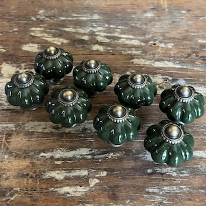 May include: A set of eight dark green ceramic cabinet knobs with a fluted, pumpkin-like shape. Each knob features a central metallic silver-toned circular cap with a decorative ridged edge, providing a contrast to the glossy dark green finish.