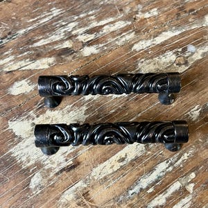 May include: Two dark metal drawer pulls featuring a cylindrical shape with a raised, swirling, embossed pattern along the center. Each handle has smooth, flat ends and is supported by two small, conical mounting posts on the underside.