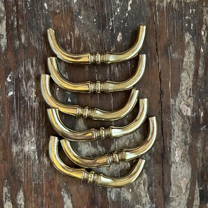 May include: A set of six curved brass drawer pulls arranged in a vertical stack. Each pull features a polished metallic finish with a central decorative ring detail and rounded ends, designed for attachment to furniture cabinetry or drawers.