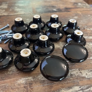 May include: A collection of twelve round, glossy black ceramic cabinet knobs with a smooth, reflective finish. Each knob features a central brass-threaded insert on the underside for mounting, accompanied by a set of silver metal mounting screws.