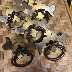 May include: A collection of four brass drawer pulls featuring ornate, decorative backplates with intricate scrollwork and leaf-like edges. Each pull includes a hinged, oval-shaped drop handle attached to the center of the metal plate.