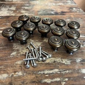 May include: A collection of twelve round cabinet knobs with a dark bronze finish and concentric circular detailing, accompanied by a set of twelve silver-colored metal mounting screws for installation on furniture or cabinetry doors and drawers.