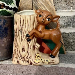 May include: A ceramic planter shaped like a brown deer standing next to a textured beige tree stump. The deer has large black eyes and black hooves, while the base of the tree stump features small yellow flowers with black centers.