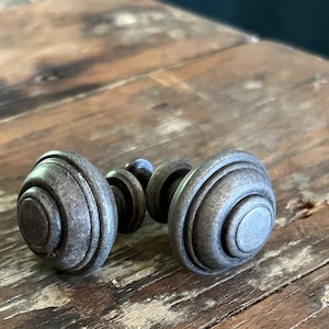 May include: Two round cabinet knobs with a dark, weathered metallic finish. Each knob features a stepped, concentric circular design with a raised center and a threaded metal bolt extending from the back for mounting purposes.