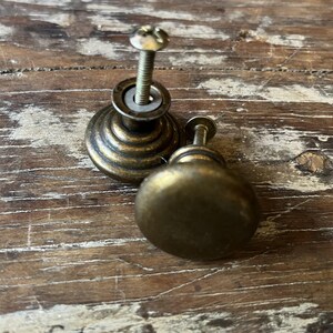 May include: Two round cabinet knobs with a dark brass finish. One knob features a stepped, tiered base design, while the other has a smooth, rounded top. Both knobs include a threaded metal screw extending from the back for installation purposes.