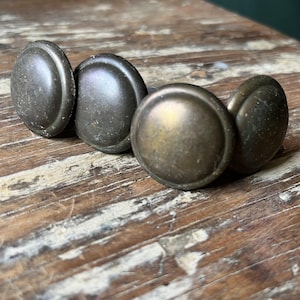 May include: Four circular metal knobs with a dark, weathered bronze finish. Each knob features a smooth, slightly domed center surrounded by a raised outer rim. The surface of the metal shows signs of age and patina with subtle variations in color.