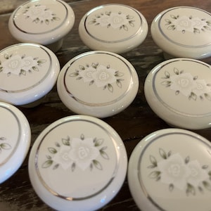 May include: A collection of eight round, off-white ceramic cabinet knobs. Each knob features a central floral design with white petals and green leaves, surrounded by a thin, circular silver metallic rim along the edge of the top surface.