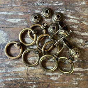 May include: A collection of antique brass-toned metal ring pulls for furniture. Each pull features a circular ring attached to a circular mounting base with a central square hole. The hardware has a weathered, metallic finish with visible screw stems.