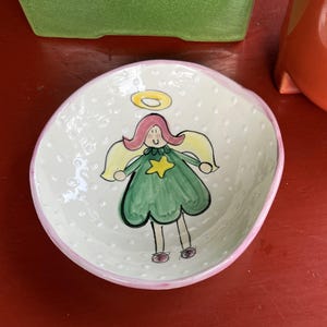 May include: A round ceramic bowl with a white interior featuring a hand-painted illustration of an angel with pink hair, a green dress, a yellow star, and a yellow halo. The bowl has a textured surface with raised dots and a pink painted rim.